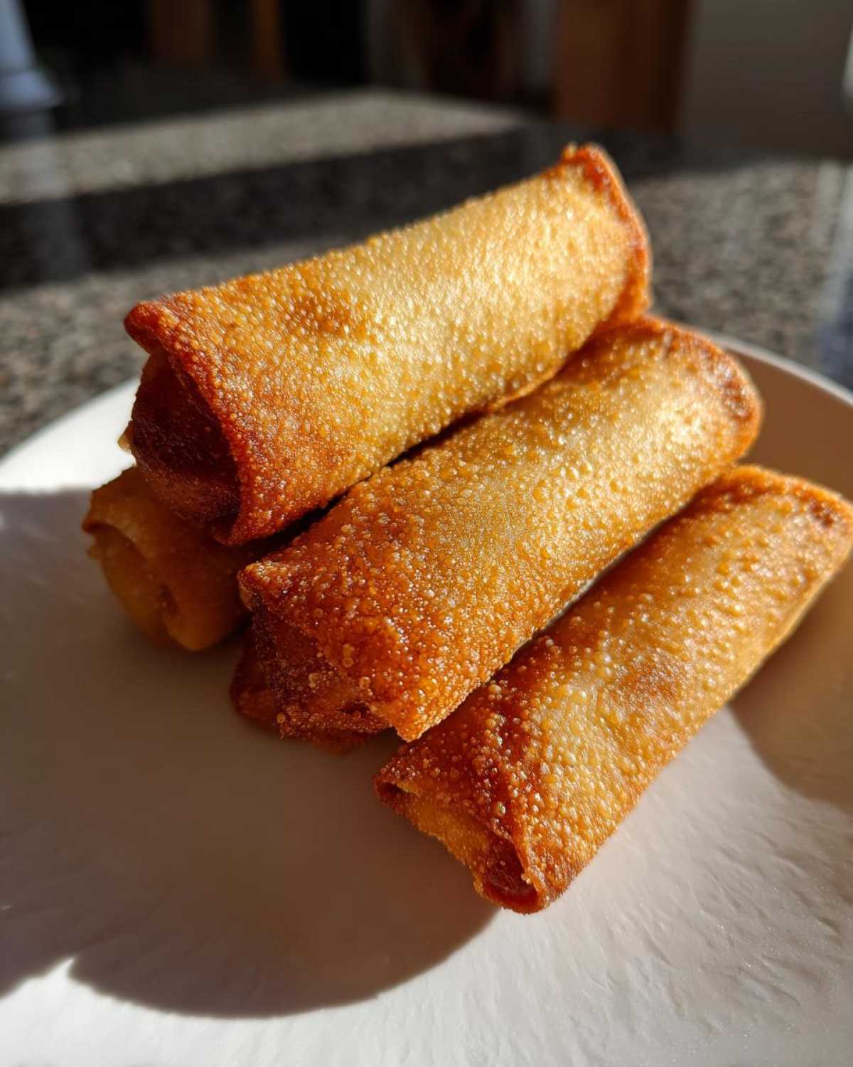 Four golden brown, crispy fried egg roll appetizers stacked on a white plate under bright sunlight.
