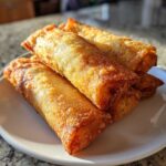 Close-up of several perfectly fried, golden brown egg roll appetizers stacked on a white plate.