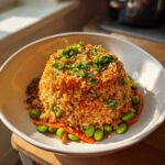 A molded serving of crispy rice salad topped with herbs and nuts, surrounded by bright green edamame and sliced carrots.
