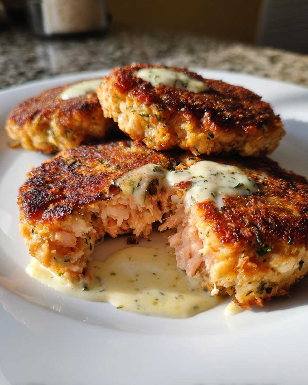 Close-up of three crispy salmon cakes, one broken open showing flaky pink salmon inside, drizzled with a creamy sauce.