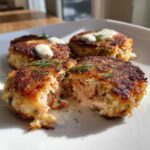 Close-up of crispy salmon cakes, one broken open showing flaky salmon interior and creamy sauce.