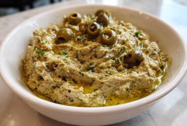 A close-up of a creamy, savory dirty martini dip topped with green olives and olive oil.