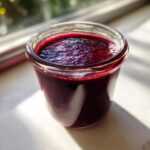 Close-up of thick, dark purple elderberry syrup recipe filling a small glass jar, highlighted by sunlight.