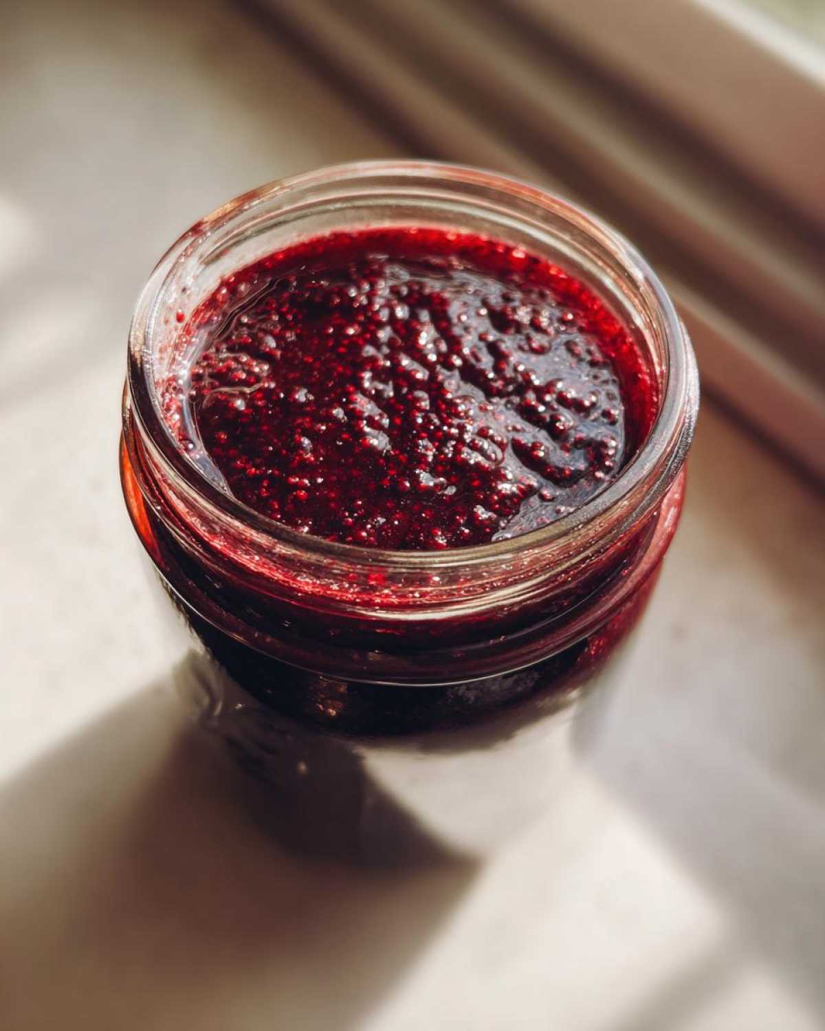Overhead view of thick, dark red elderberry syrup recipe mixture in a clear glass jar, catching sunlight.