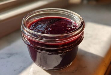 Close-up of rich, dark purple elderberry syrup recipe filling a small glass jar, catching sunlight.