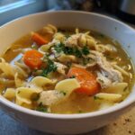 Close-up of a white bowl filled with steaming chicken noodle soup, featuring egg noodles, shredded chicken, carrots, and parsley.