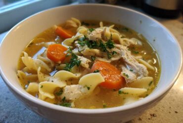 Close-up of a white bowl filled with steaming chicken noodle soup, featuring egg noodles, shredded chicken, carrots, and parsley.