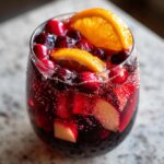 Close-up of a glass filled with bubbling, deep red holiday sangria, garnished with cranberries, apple chunks, and orange slices.