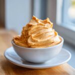 A close-up of perfectly piped, fluffy orange pumpkin fluff served in a small white bowl on a saucer.