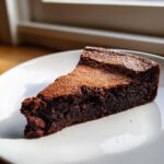 A rich, dense slice of flourless chocolate torte dusted with cocoa powder on a white plate.