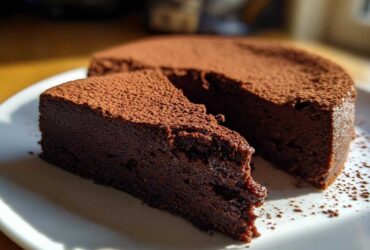 A slice of dense, dark flourless chocolate torte dusted heavily with cocoa powder, sitting next to the main cake.