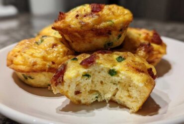 A close-up of several fluffy, golden brown egg bites with bacon and green onions, one of which is bitten into, revealing the texture of the egg bites.