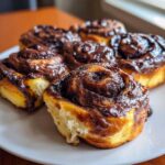 Close-up of fluffy chocolate cinnamon rolls topped with a thick, glossy chocolate glaze on a white plate.