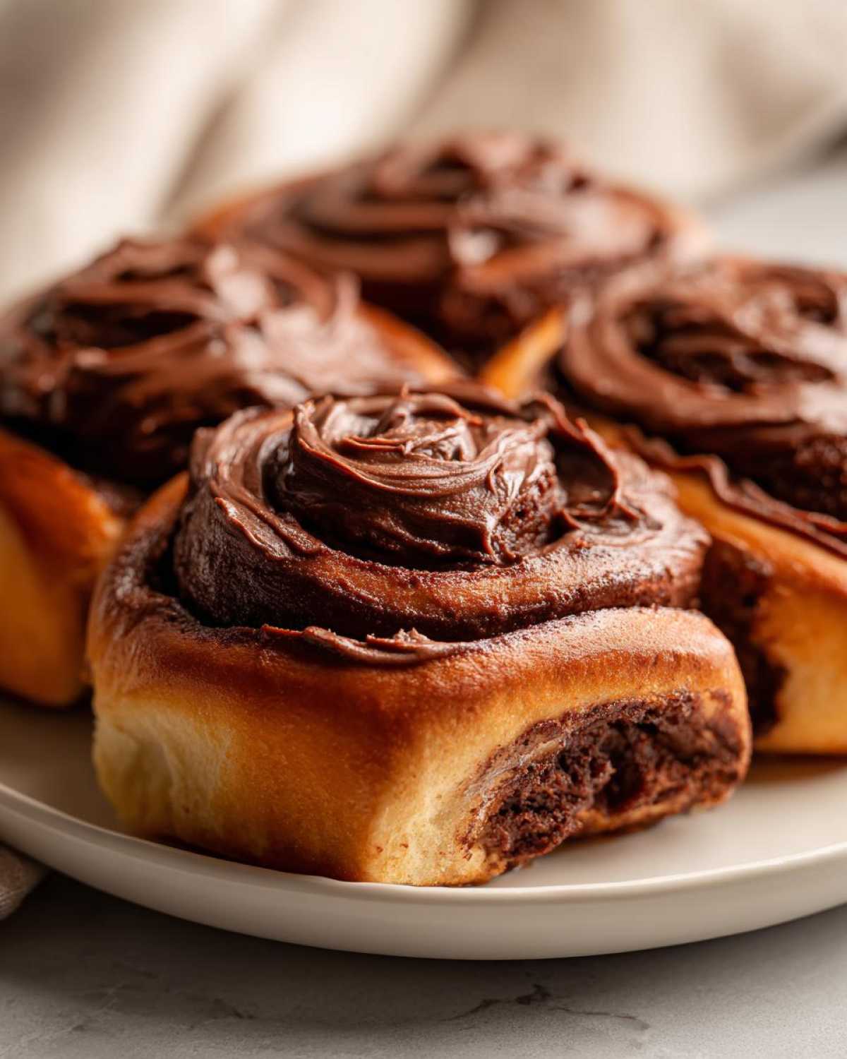 Close-up of several fluffy chocolate cinnamon rolls topped with rich, dark chocolate frosting on a white plate.