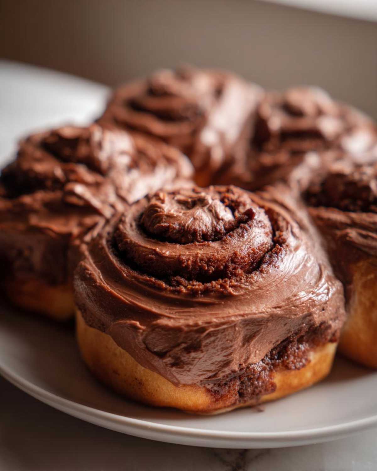 Close-up of several fluffy chocolate cinnamon rolls generously topped with rich chocolate frosting on a white plate.