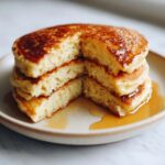 Close-up of a stack of fluffy greek yogurt pancakes with a wedge cut out, drizzled generously with syrup.
