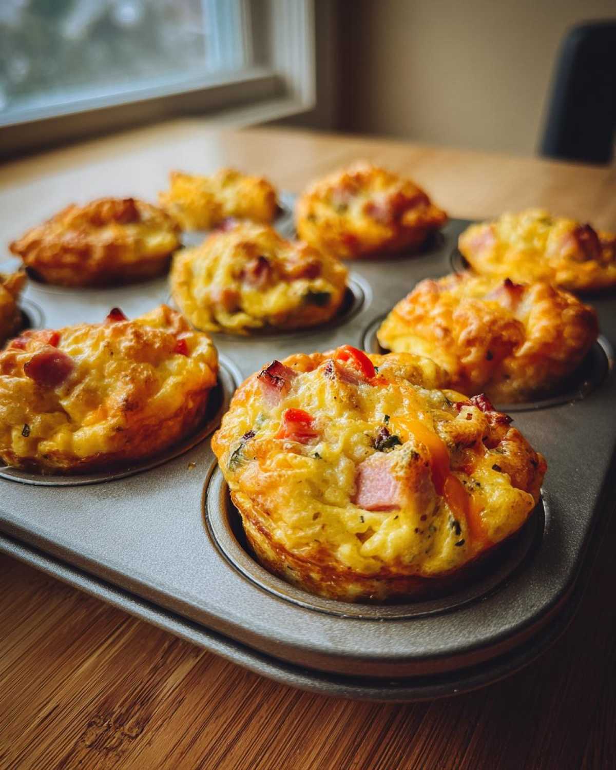 Close-up of several golden brown muffin tin egg bites filled with ham and peppers, resting in a metal baking pan.
