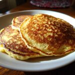 A stack of three golden-brown, fluffy oatmeal pancakes served on a white plate, highlighted by natural window light.