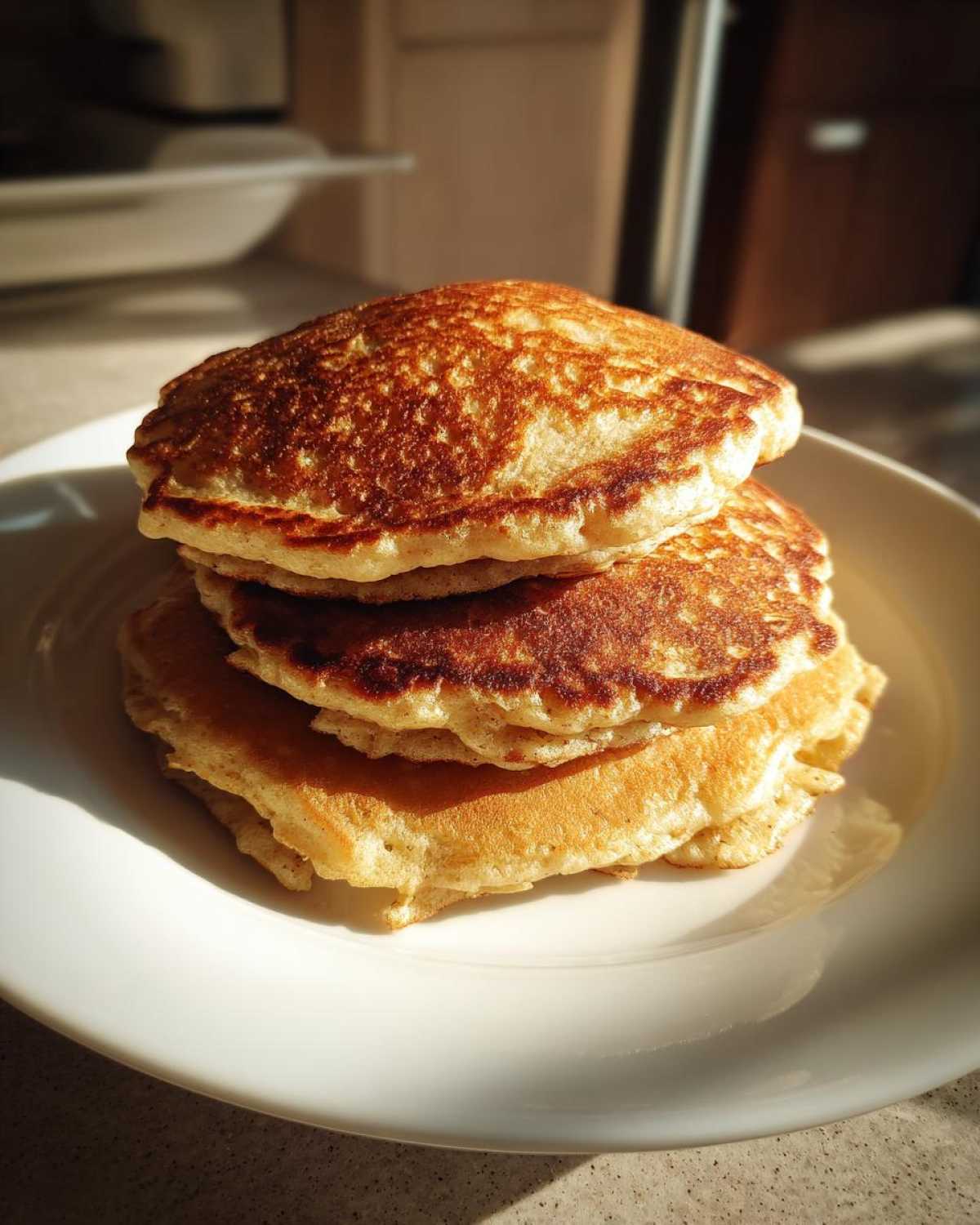 A stack of three golden brown, fluffy oatmeal pancakes sitting on a white plate, highlighted by bright sunlight.