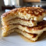 Close-up of a stack of three incredibly fluffy pancakes cut in half, showing the airy interior and drizzled with syrup.