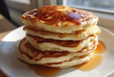 A tall stack of four fluffy sourdough discard pancakes drizzled generously with maple syrup on a white plate.