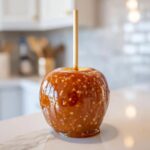 A glossy, finished caramel apple with a wooden stick inserted, sitting on a white marble counter.