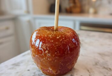 A single, glossy red caramel apple on a stick, sprinkled with sugar crystals, sitting on a marble counter.