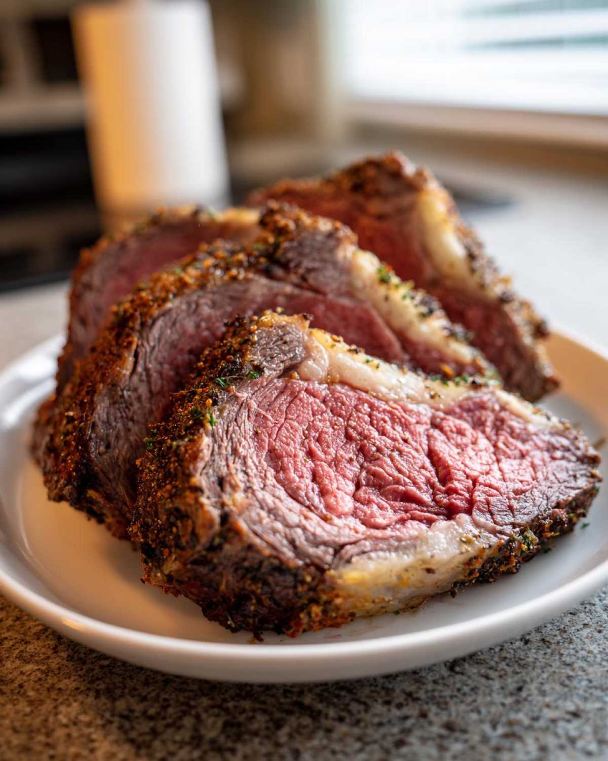 Close-up of three thick slices of medium-rare prime rib recipe with a dark, herbed crust on a white plate.