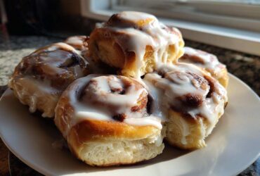 A stack of warm, gooey cinnamon roll pastries generously topped with dripping white icing, sitting on a white plate.