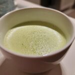 Close-up of a frothy, light green beverage, possibly a creamy celery soup, served in a white bowl.