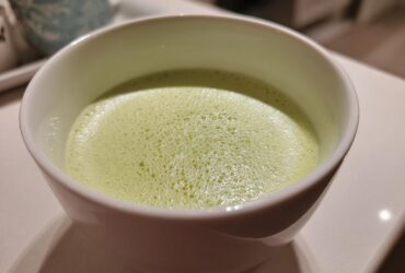 Close-up of a frothy, light green beverage, possibly a creamy celery soup, served in a white bowl.