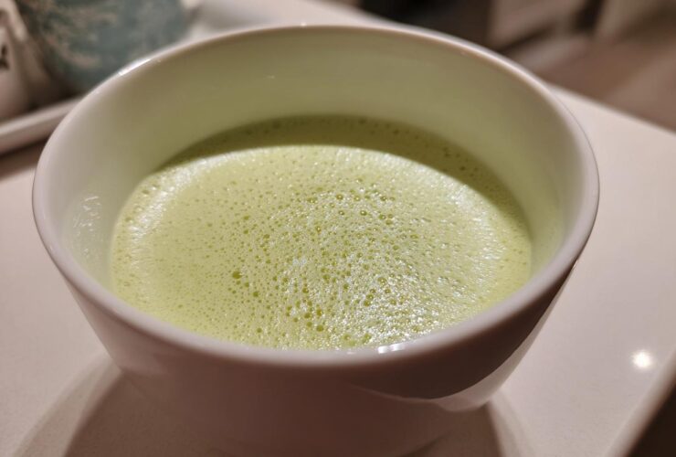 Close-up of a frothy, light green beverage, possibly a creamy celery soup, served in a white bowl.