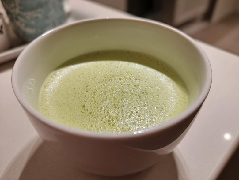 Close-up of a frothy, light green beverage, possibly a creamy celery soup, served in a white bowl.