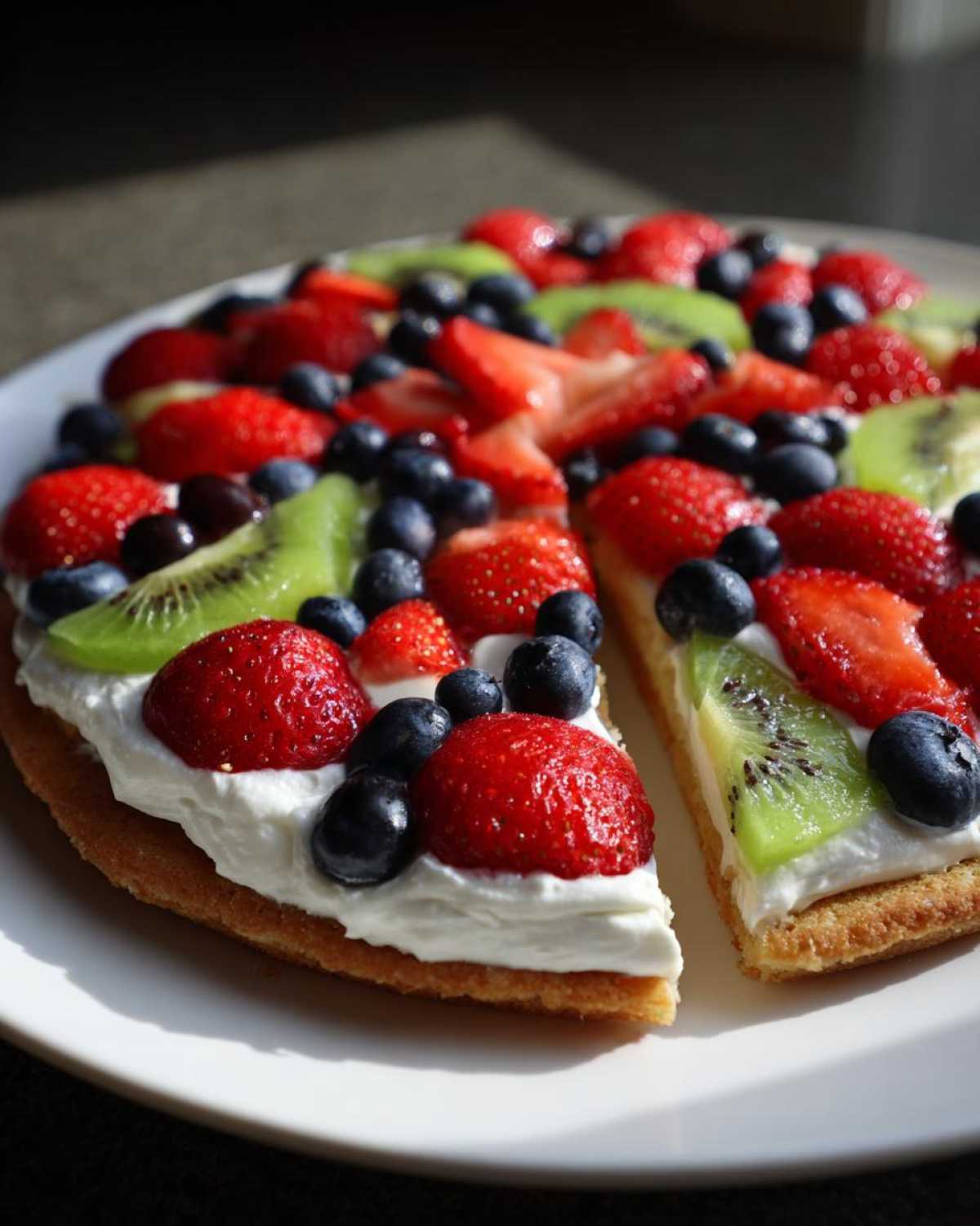 A close-up of a fruit pizza sugar cookie topped with whipped cream, strawberries, blueberries, and kiwi slices.
