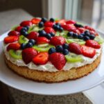 A vibrant fruit pizza sugar cookie topped with whipped cream, strawberries, kiwi slices, and blueberries.