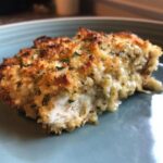 A close-up of a juicy slice of Garlic parmesan chicken bake with a golden, crispy, cheesy topping and visible white chicken meat.