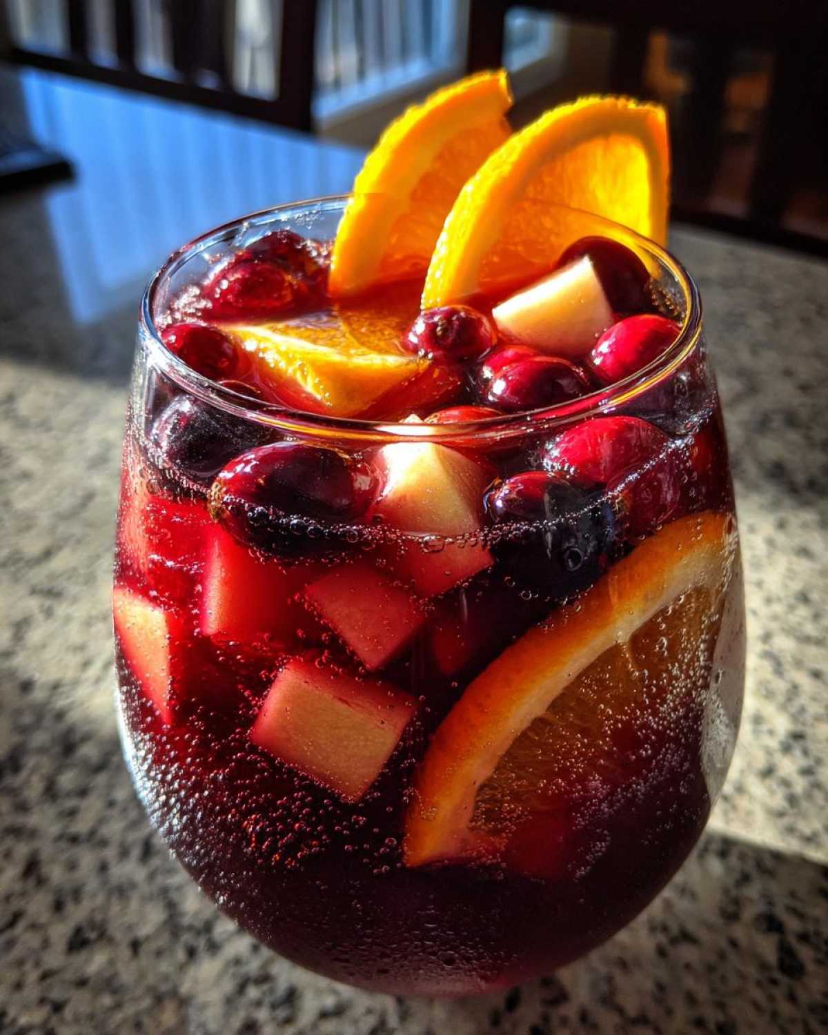 Close-up of a glass filled with bubbling red holiday sangria, garnished with orange slices, cranberries, and apple chunks.