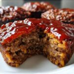 Close-up of a delicious mini meatloaf with a bite taken out, showing the moist interior and thick, shiny ketchup glaze.