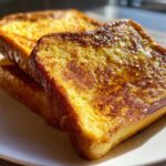 Close-up of three glistening slices of golden brown french toast stacked on a white plate.