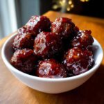 A white bowl filled with shiny, dark glazed grape jelly meatballs sitting on a wooden surface.