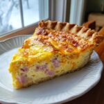 A perfect slice of ham and cheese quiche with a golden, bubbly top, served on a white plate.