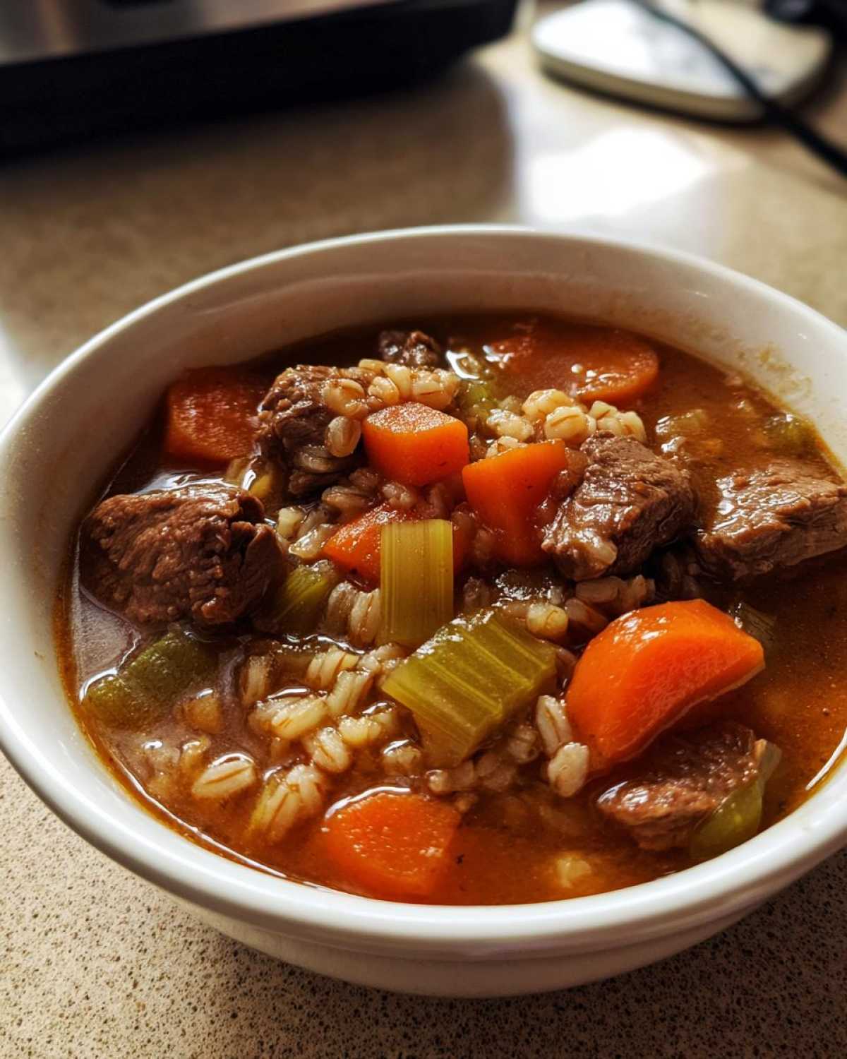 Close-up of a white bowl filled with rich beef barley soup, featuring chunks of beef, carrots, celery, and barley.