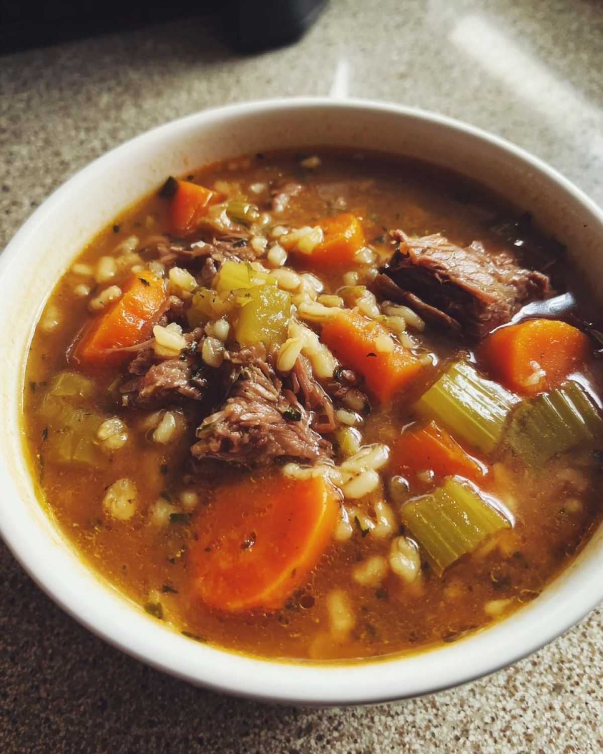 Close-up of a white bowl filled with rich beef barley soup, featuring shredded beef, large carrot chunks, and celery.