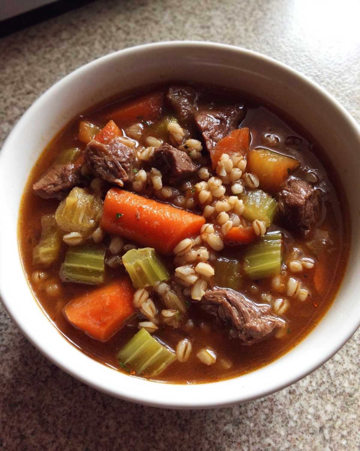 Close-up of a white bowl filled with rich beef barley soup, featuring chunks of beef, carrots, celery, and barley.