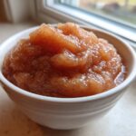 Close-up of chunky, spiced applesauce served in a small white bowl near a window.