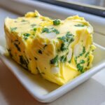 A block of homemade garlic butter mixed with chopped parsley and sliced almonds sitting on a small white dish.