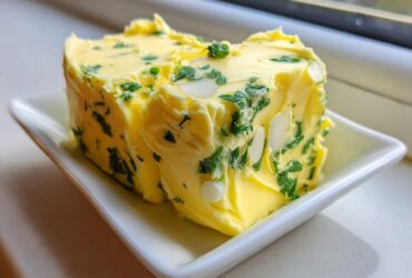 A block of homemade garlic butter mixed with chopped parsley and sliced almonds sitting on a small white dish.