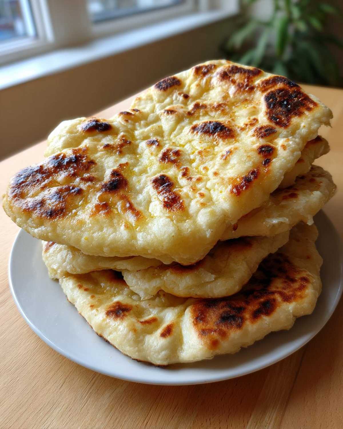 A stack of four freshly cooked homemade naan bread rounds with golden-brown bubbles and char marks, resting on a white plate.
