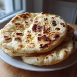 A stack of fluffy, slightly charred homemade naan bread glistening with butter on a white plate.