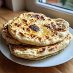 A stack of freshly made, fluffy homemade naan bread brushed with melted butter, showing charred spots.